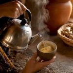 Des mains remuent doucement une tasse de thé à la maca, laissant s’échapper une vapeur délicate.